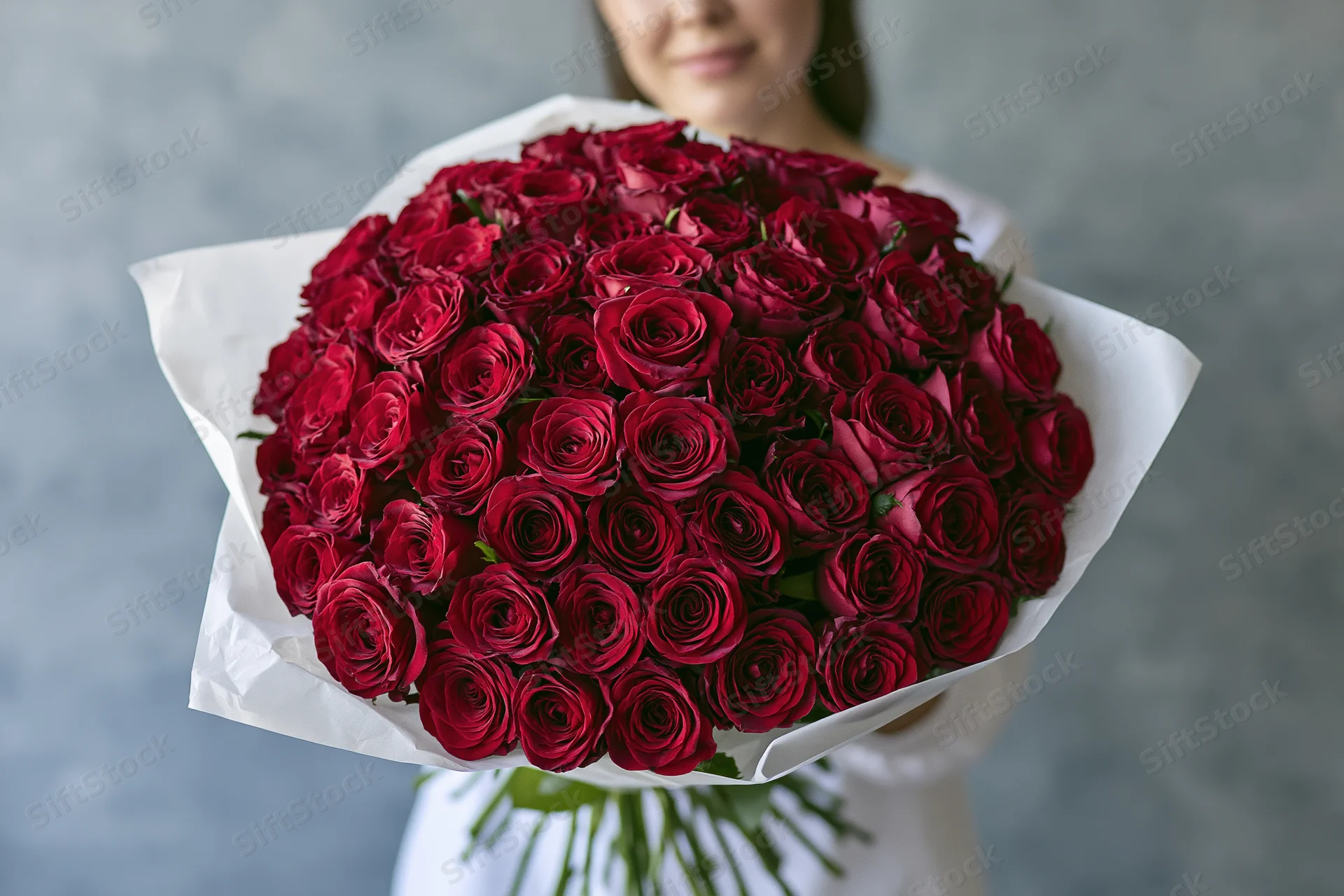 Woman holding large bouquet of deep red roses wrapped in white paper against gray background