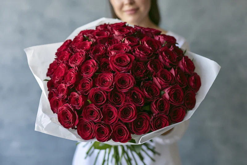 Woman holding large bouquet of deep red roses wrapped in white paper against gray background