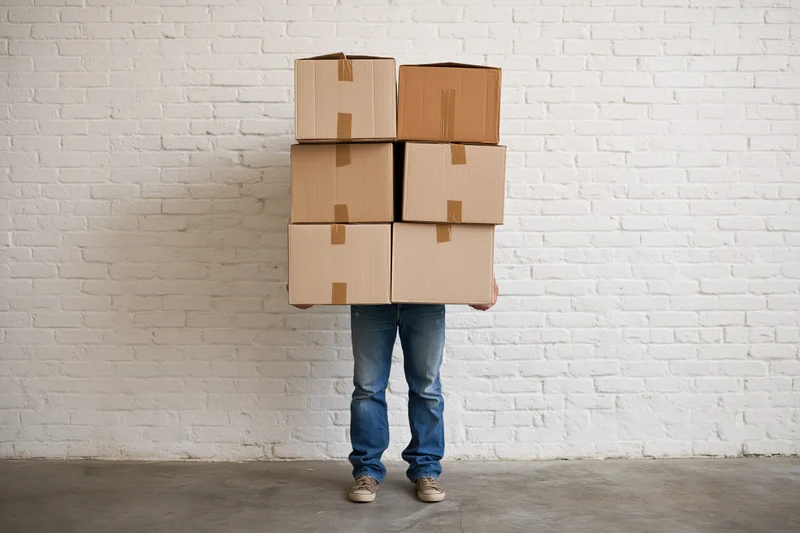 Person carrying six stacked cardboard boxes against white brick wall