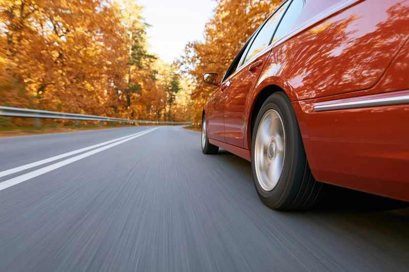 Red-orange sedan driving on a two-lane highway through an autumn forest