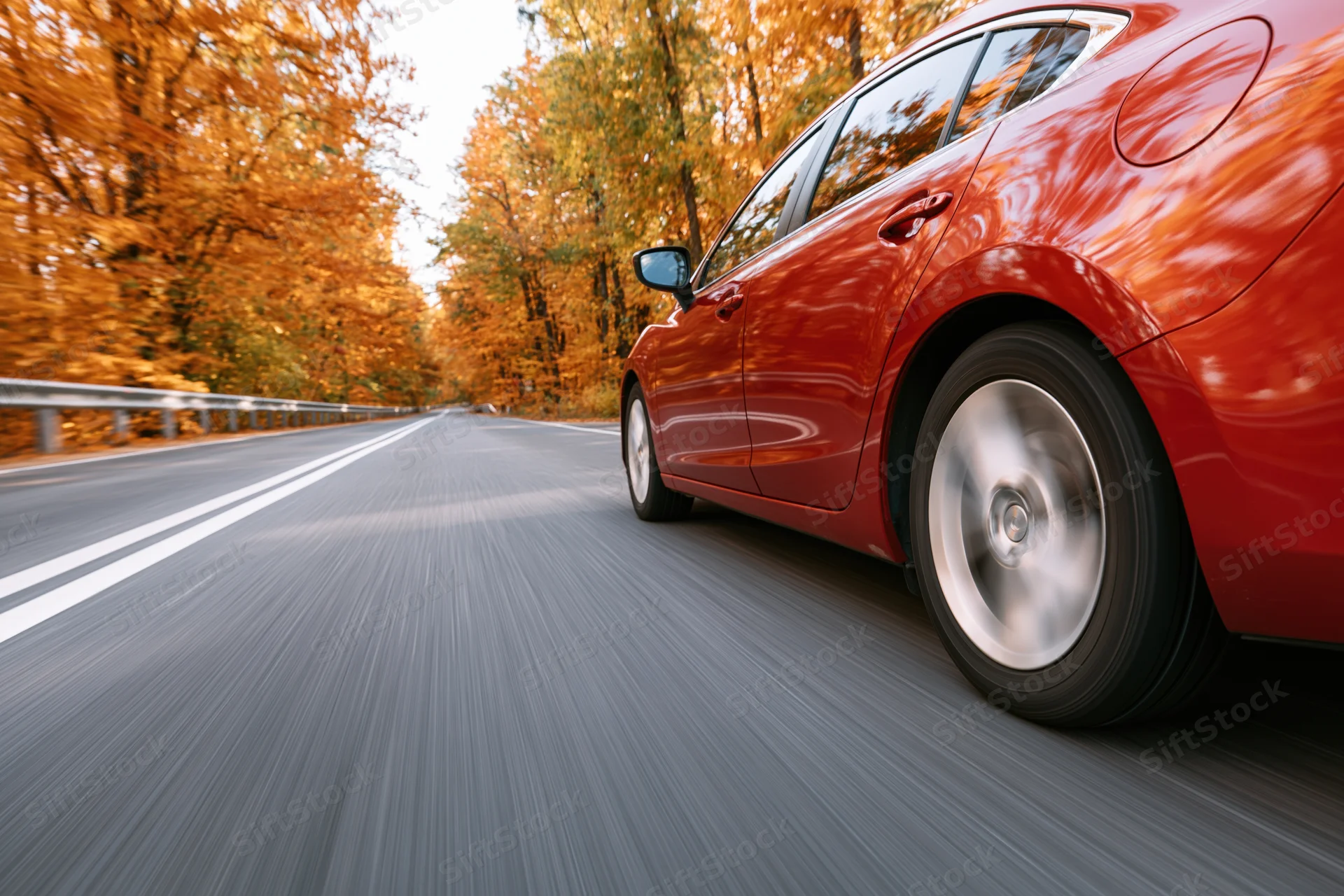 Red-orange sedan driving on a two-lane highway through autumn trees