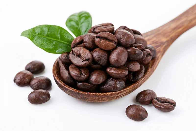 Roasted coffee beans piled in a wooden spoon with green leaves on white background