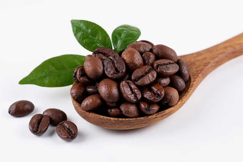 Roasted coffee beans in a wooden spoon with green leaves on white background