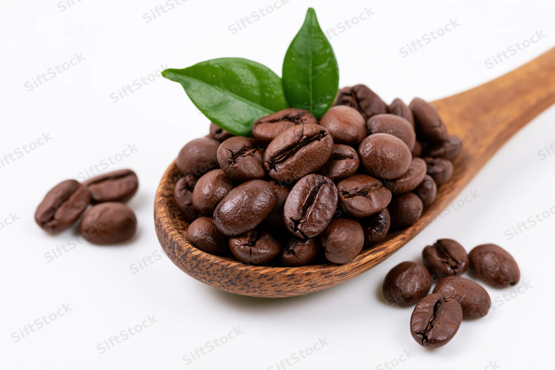 Roasted coffee beans in a wooden spoon with a green leaf on white background