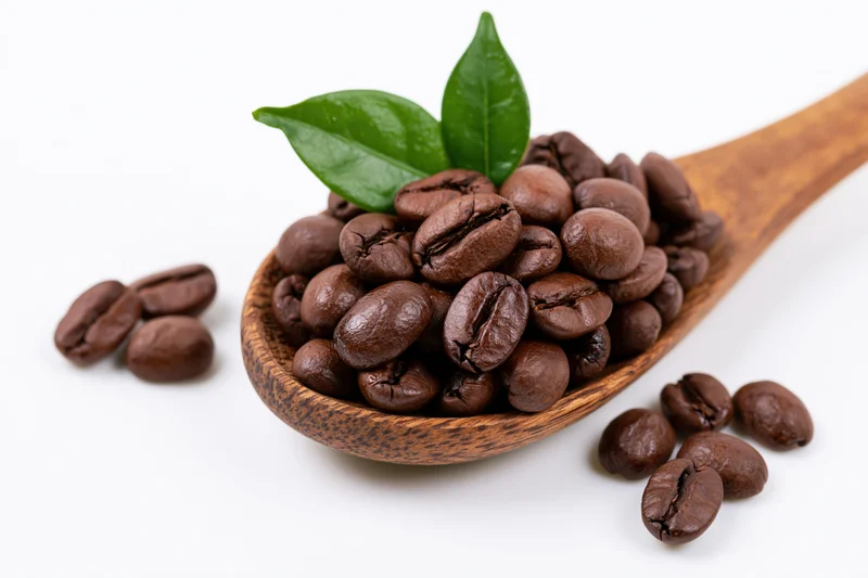 Roasted coffee beans in a wooden spoon with a green leaf on white background