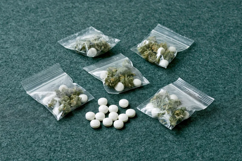 Cannabis buds and white pills in clear zip-lock bags on dark green surface