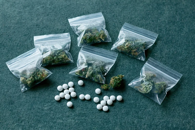Dried cannabis buds in clear zip-lock bags with white pills arranged on dark textured surface