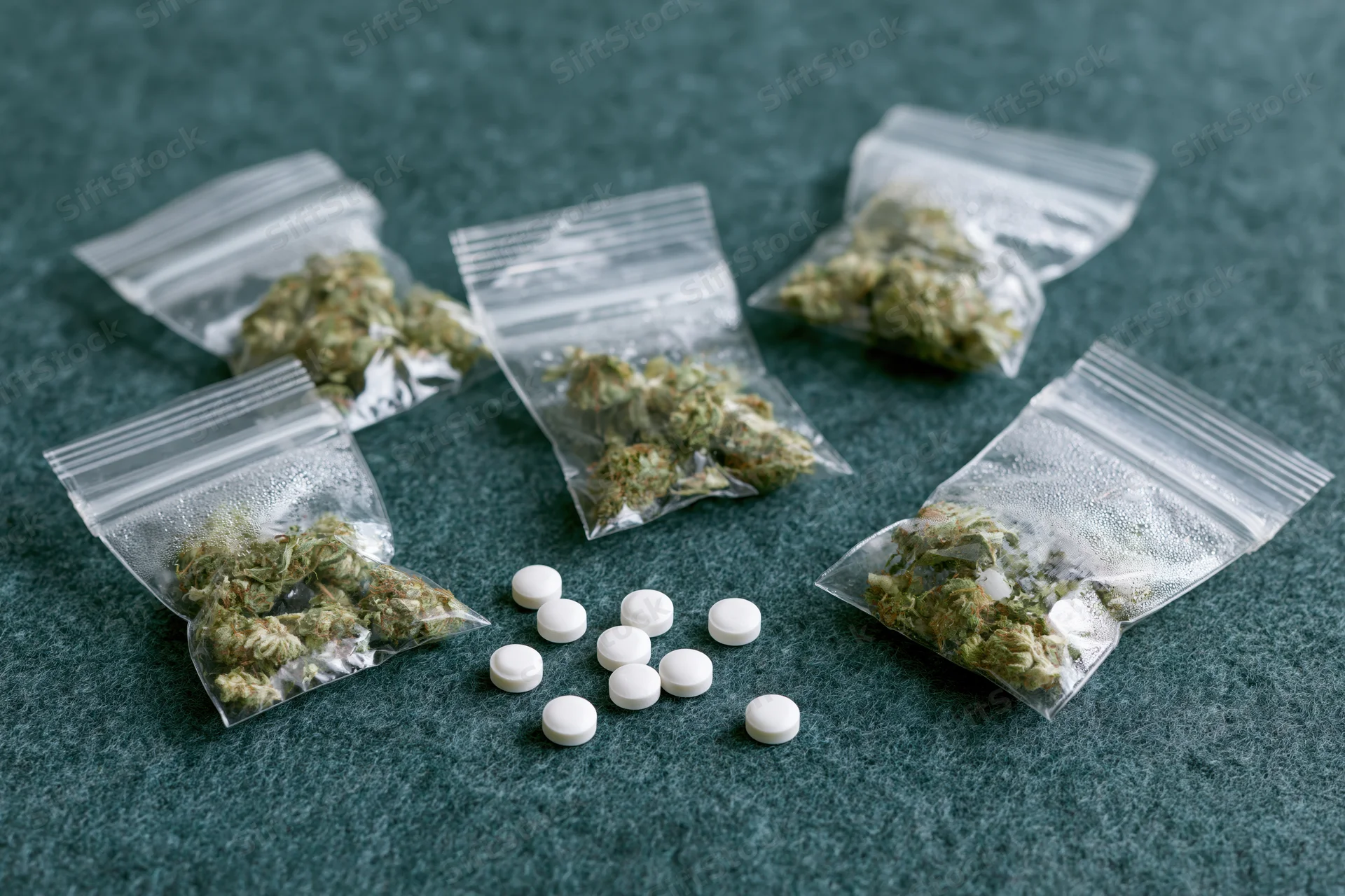 Dried cannabis buds in clear zip-lock bags with white pills arranged on a dark teal surface