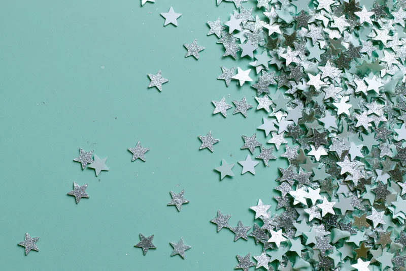 Silver and white star-shaped glitter confetti scattered across a mint green surface with copyspace on the left