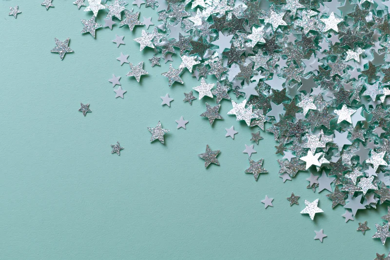 Silver and white star-shaped glitter confetti scattered across a mint green surface with copyspace on the left