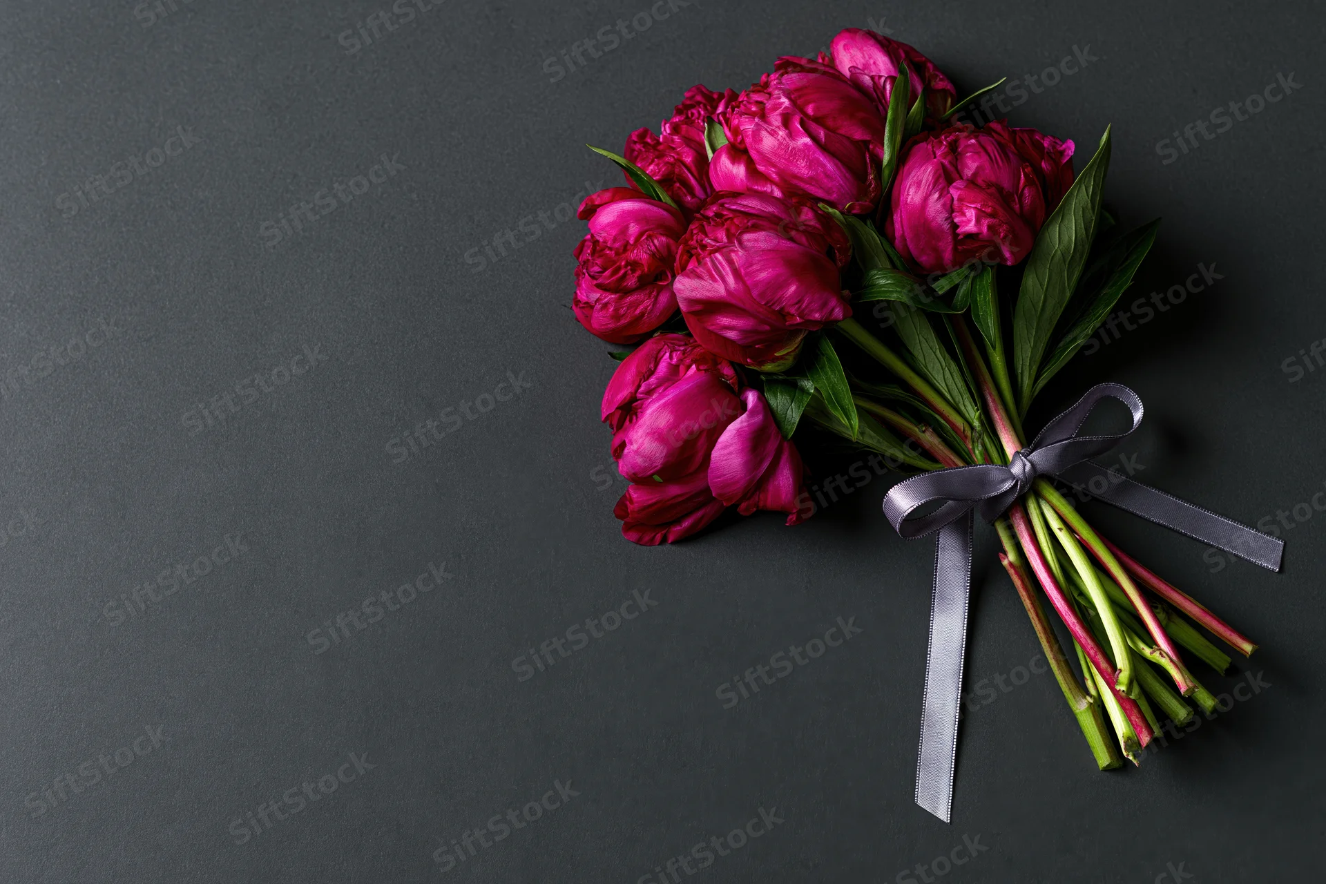 Small bouquet of deep crimson peonies with green stems and leaves tied with a dark ribbon on a black background