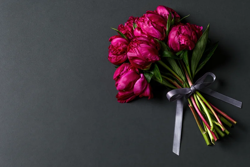 Small bouquet of deep crimson peonies with green stems and leaves tied with a dark ribbon on a black background