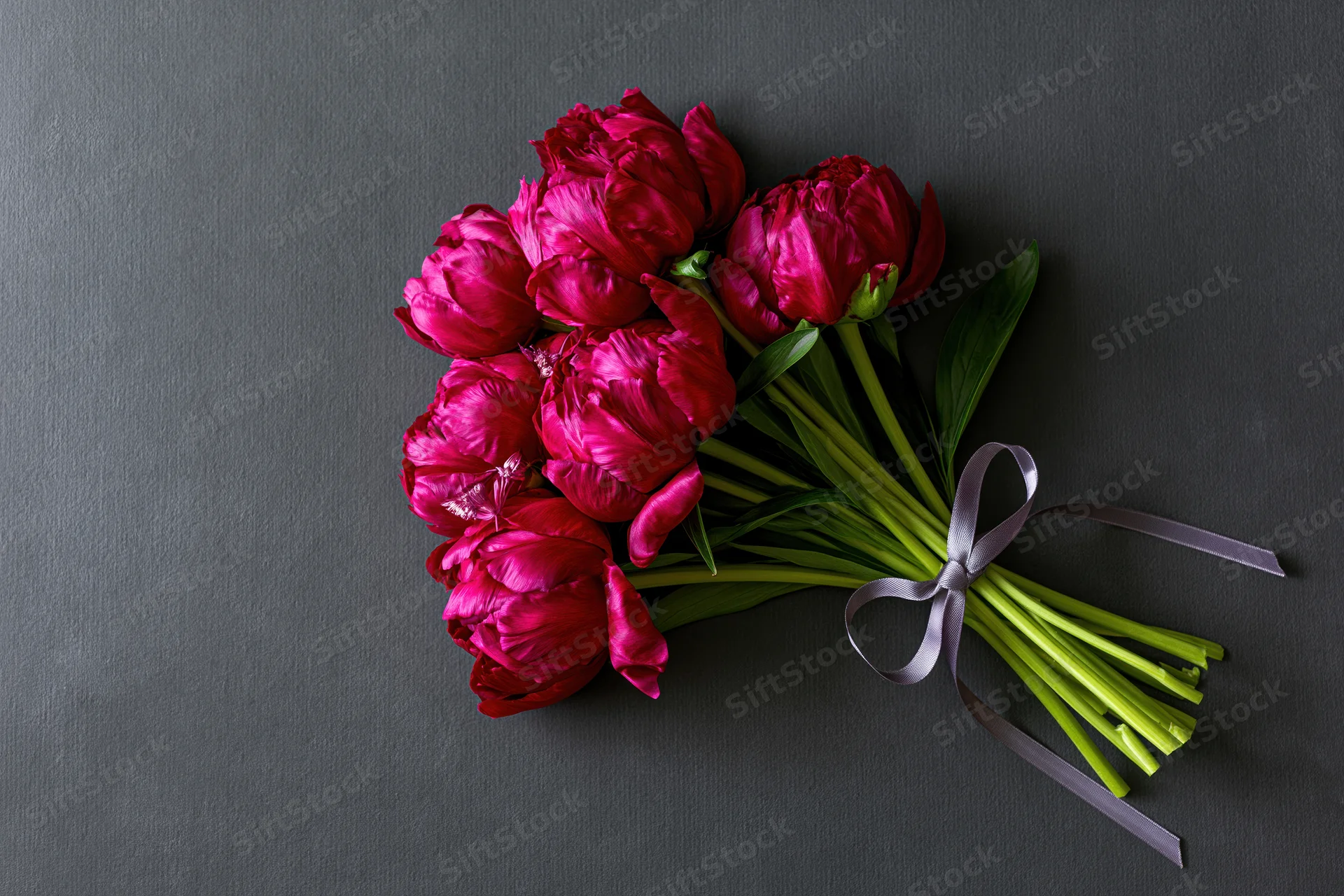 Small bouquet of deep crimson peonies with green stems and leaves tied with a ribbon on a dark gray surface