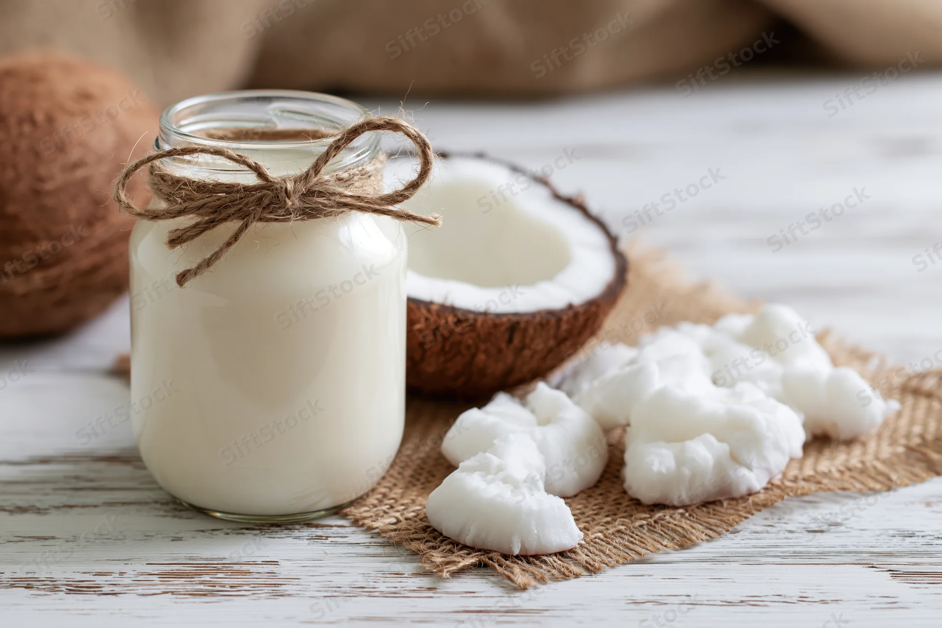 Glass jar of coconut milk tied with twine on a white wooden table beside coconut halves and shredded coconut meat on burlap
