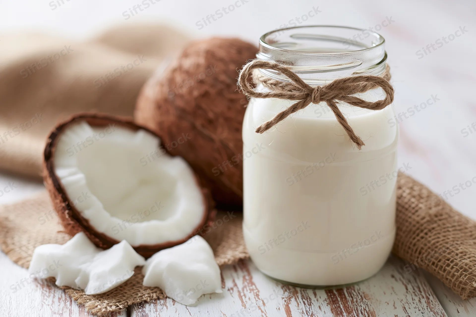 Glass jar of coconut milk with twine bow beside halved coconut and coconut pieces on burlap cloth