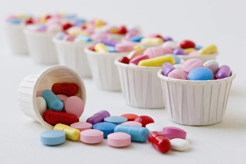 Colorful pills and capsules scattered from small white paper cups on a white surface