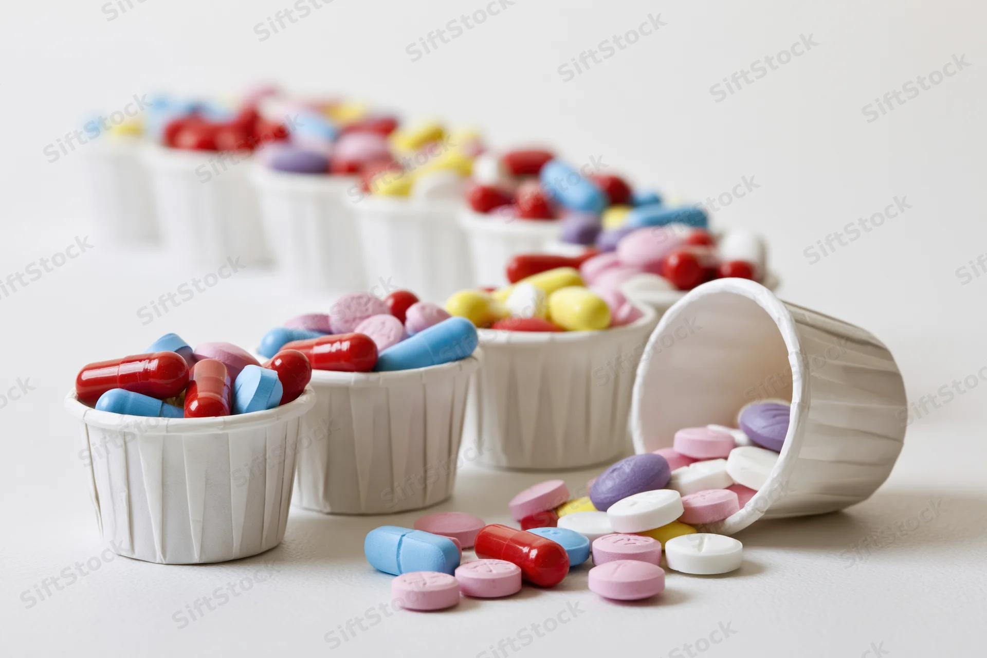 Colorful pills and capsules spilled from white paper medicine cups on a plain surface