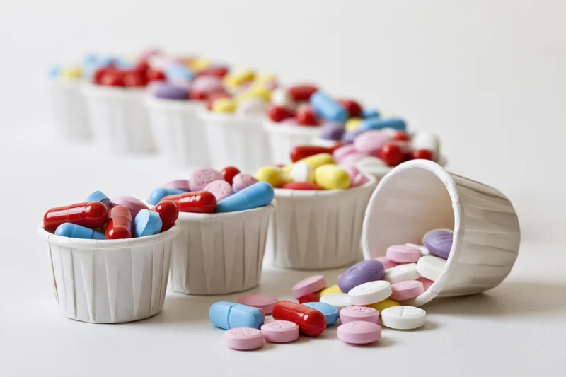 Colorful pills and capsules spilled from white paper medicine cups on a plain surface