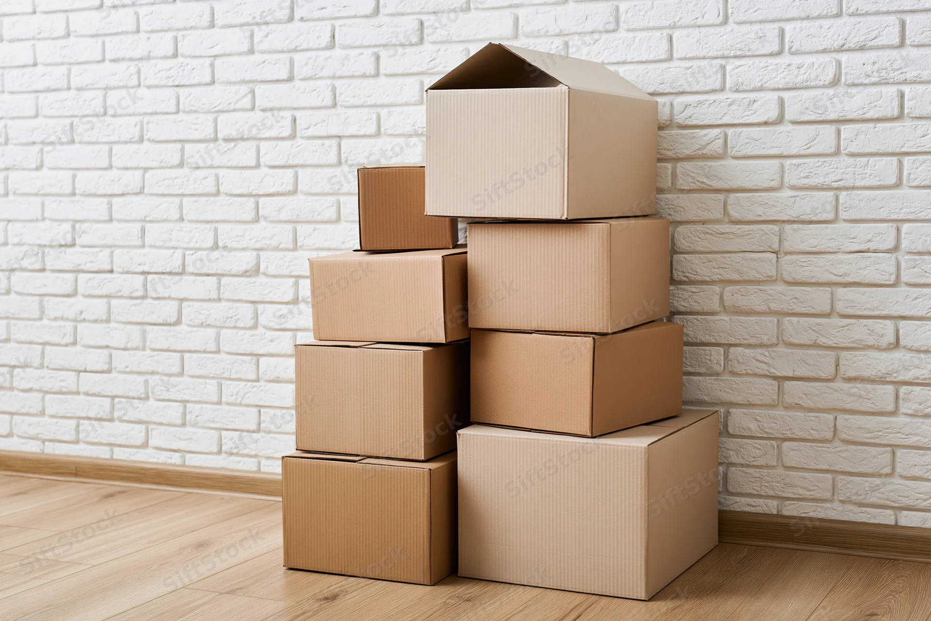 Stack of cardboard moving boxes piled against a white brick wall in a room with wooden floor
