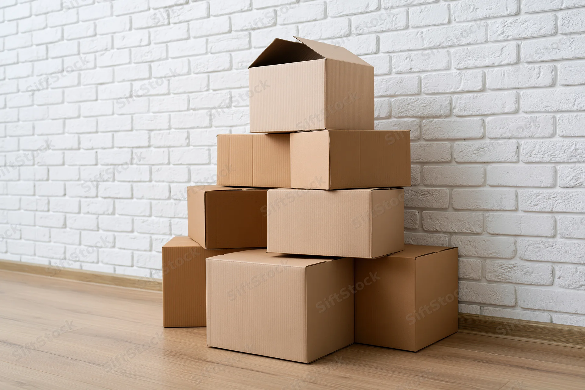 Stack of cardboard moving boxes piled against a white brick wall on a wooden floor