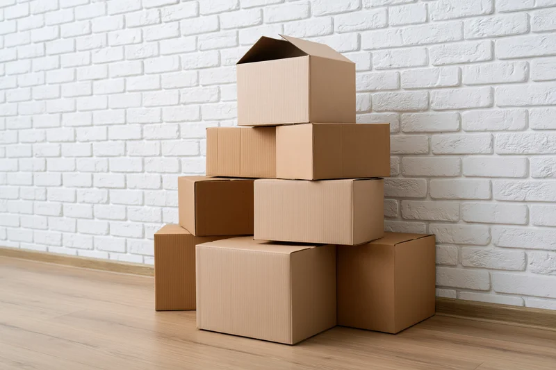 Stack of cardboard moving boxes piled against a white brick wall on a wooden floor