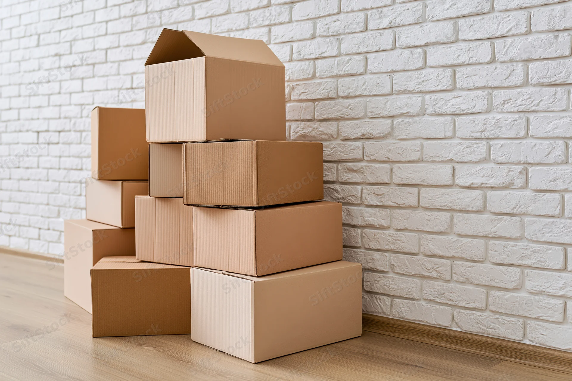 Stack of cardboard moving boxes piled against a white brick wall in a room with wooden flooring