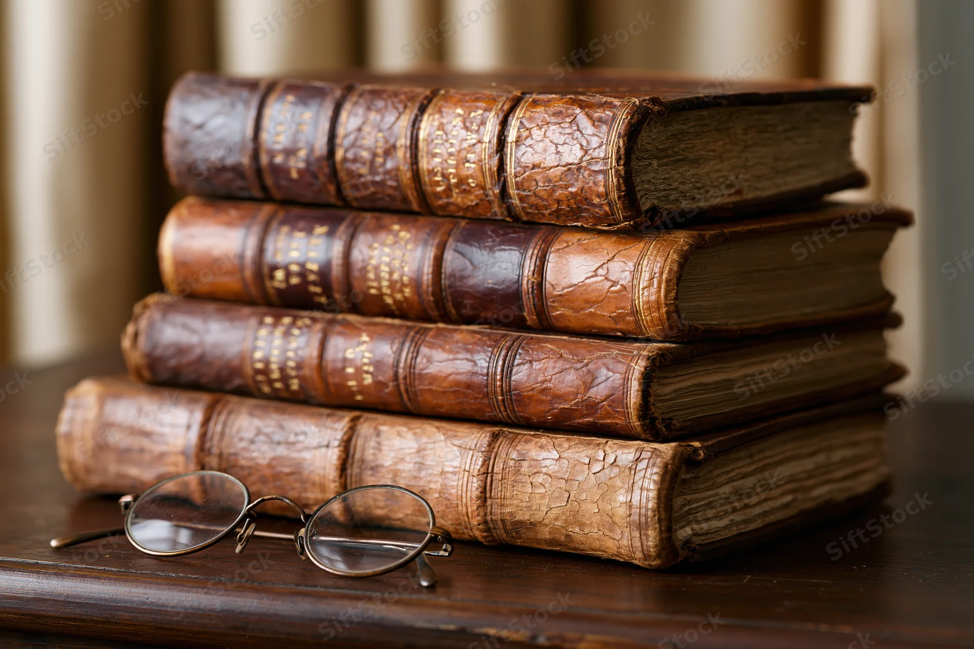 Stack of four antique leather-bound books on wooden table with eyeglasses beside them