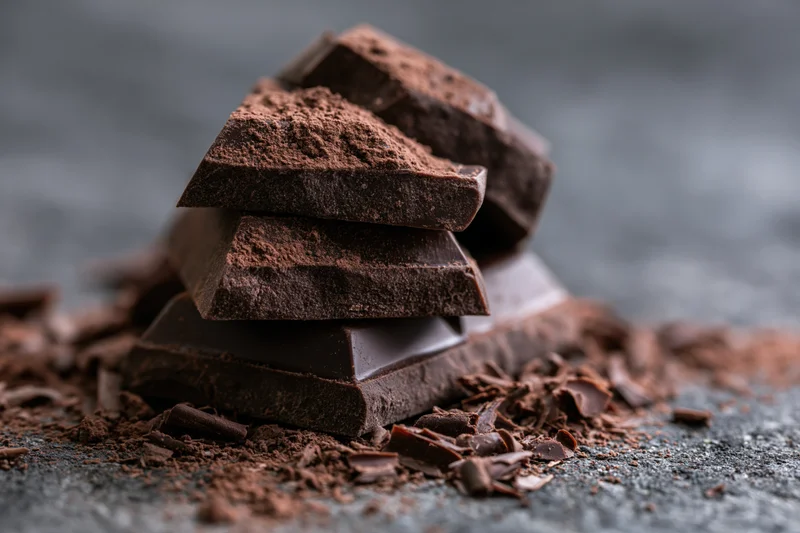 Broken dark chocolate chunks stacked and dusted with cocoa powder on a dark surface