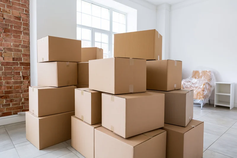 Stacks of cardboard moving boxes arranged in a bright empty apartment with exposed brick wall and large window