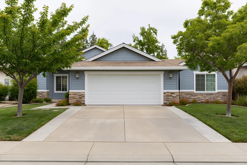 Suburban single-story house with white two-car garage and concrete driveway flanked by green lawn and trees