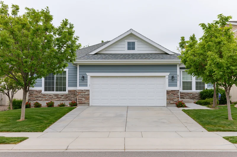 Suburban single-story house with white two-car garage and concrete driveway surrounded by green lawn and trees