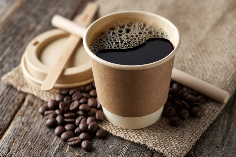 Takeaway paper coffee cup filled with black coffee and foam on burlap over wooden table with coffee beans and stirrer