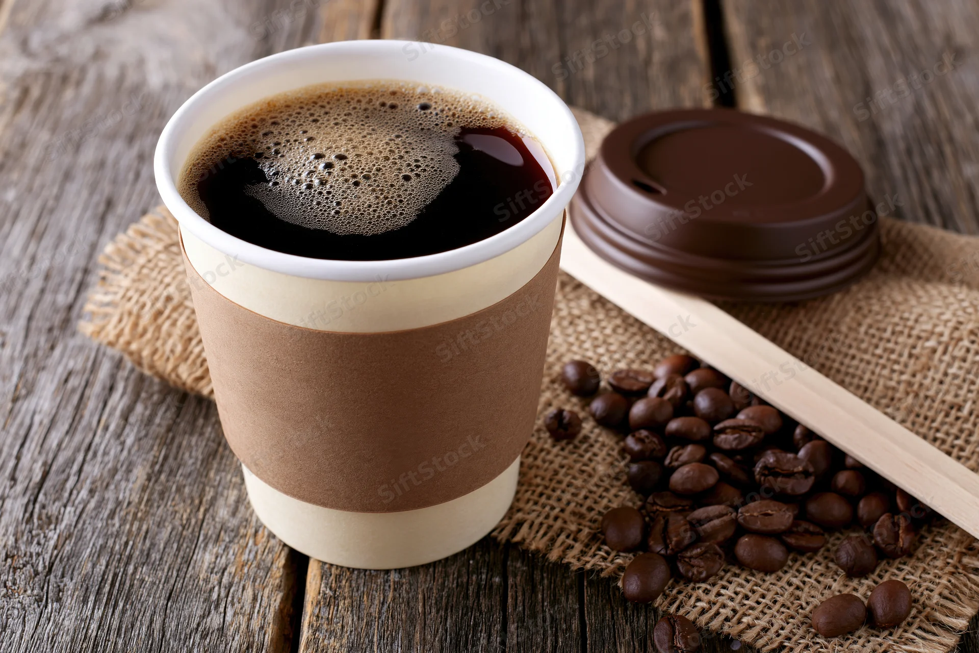 Takeaway paper coffee cup filled with black coffee on wooden table with coffee beans and burlap cloth