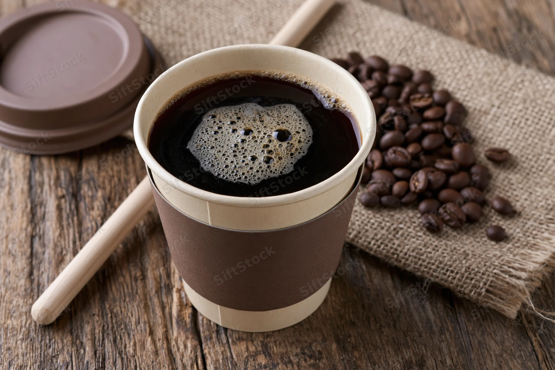 Takeaway paper coffee cup filled with black coffee and foam on wooden table with coffee beans and burlap cloth