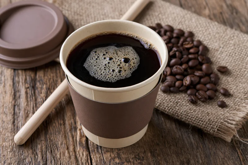 Takeaway paper coffee cup filled with black coffee and foam on wooden table with coffee beans and burlap cloth