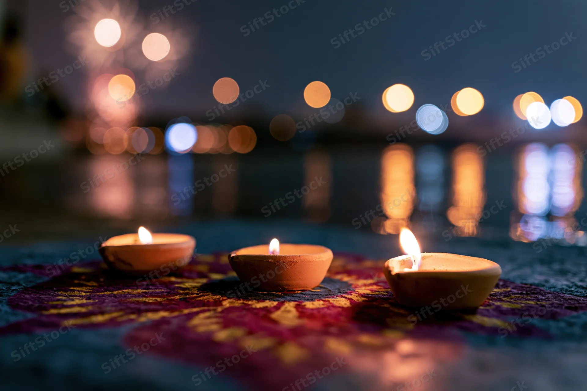 Three clay diya oil lamps burning with small flames on a colorful patterned surface near water at night