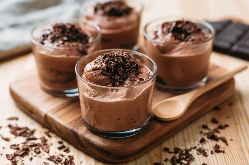 Chocolate mousse in clear glasses topped with chocolate shavings on a wooden board with a spoon nearby