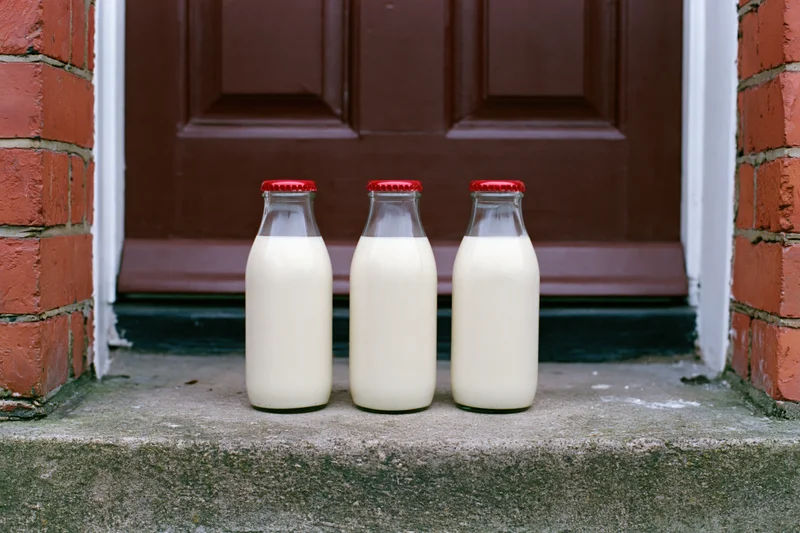 Three glass milk bottles with red foil caps on a concrete doorstep in front of a brown door