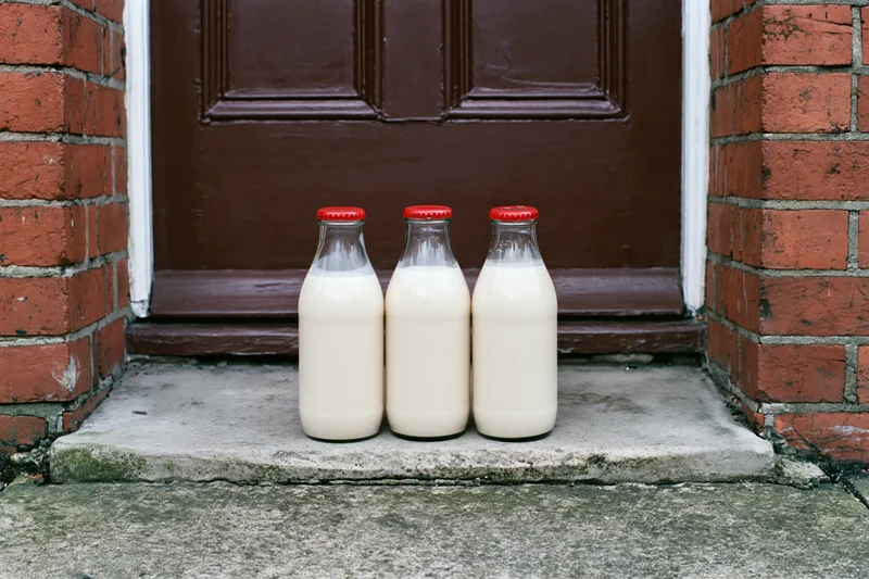 Three glass milk bottles with red foil caps on a doorstep in front of a brown door and brick wall