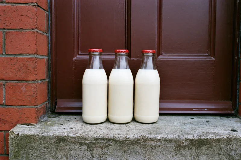Three glass milk bottles with red foil caps on a concrete doorstep in front of a brown door and red brick wall