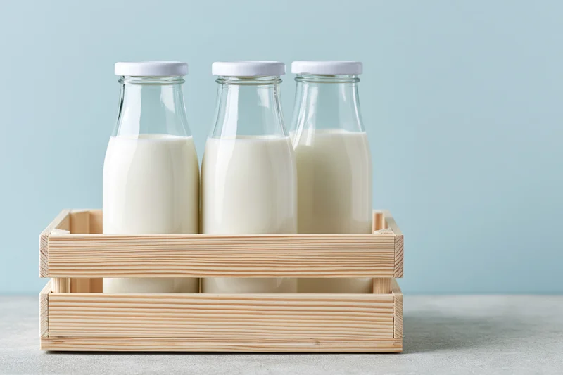 Three glass milk bottles with white caps arranged in a wooden tray on a light blue background
