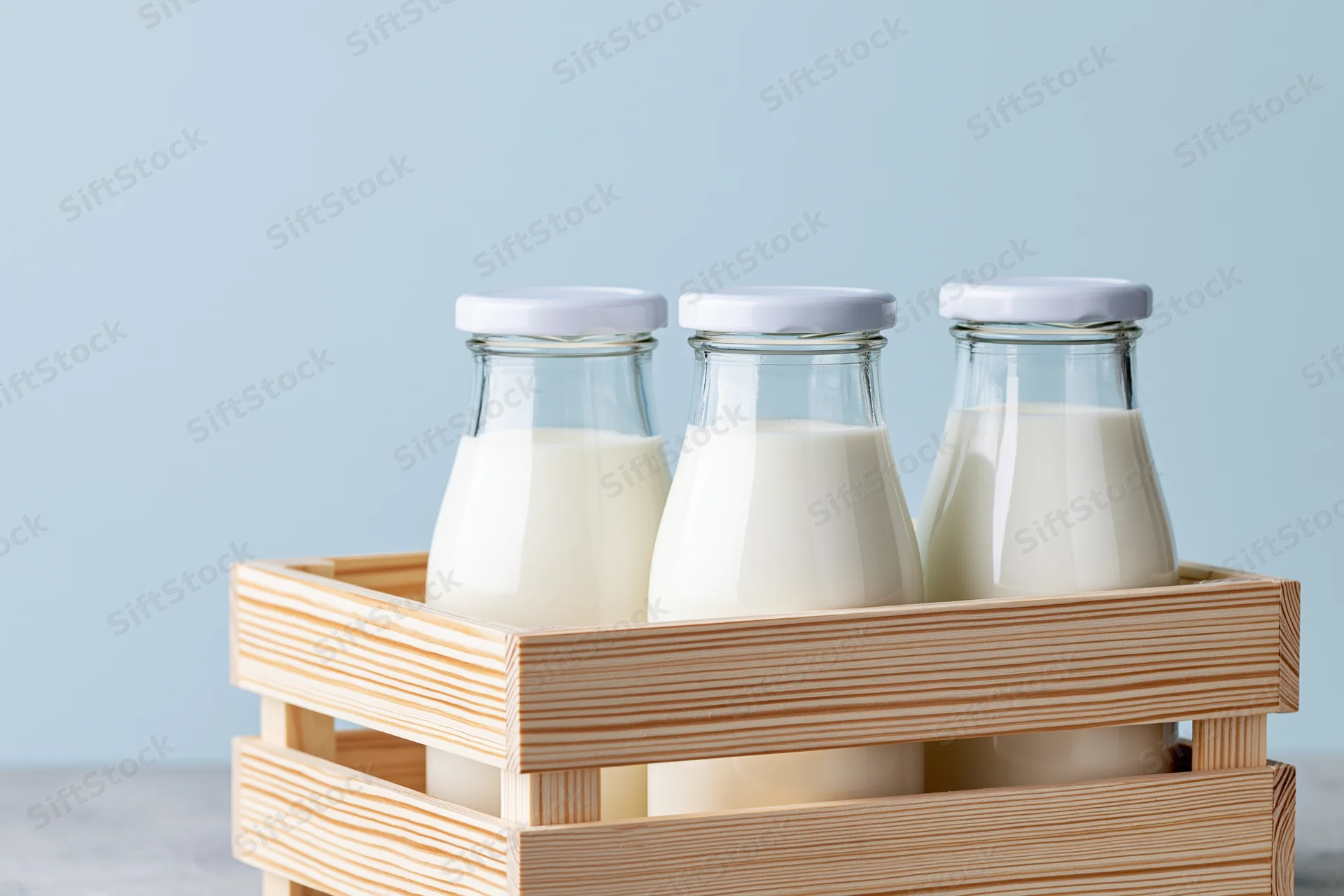 Three glass milk bottles with white caps arranged in a wooden crate on a light blue background