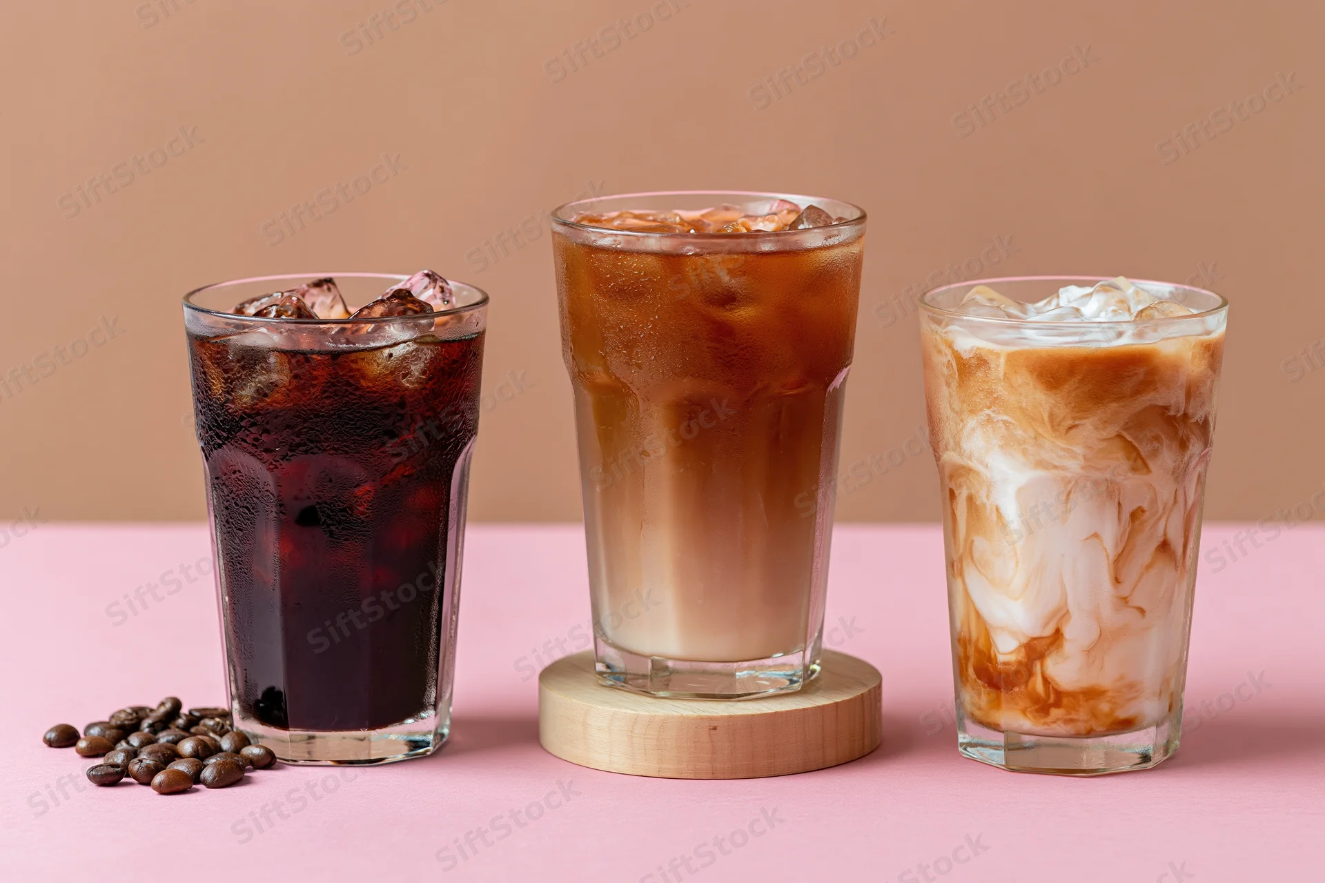 Three iced coffee drinks arranged side by side on a pink surface with coffee beans and wooden coaster