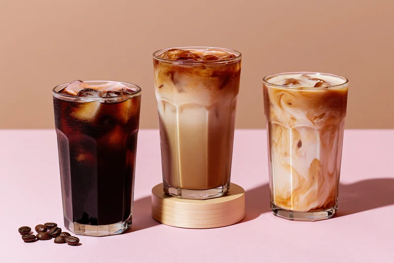 Three iced coffee drinks with cream swirls and ice cubes on a pink surface