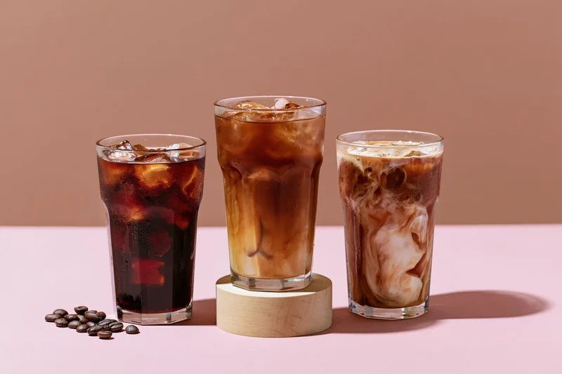 Three iced coffee drinks with ice and coffee beans arranged on a pink surface