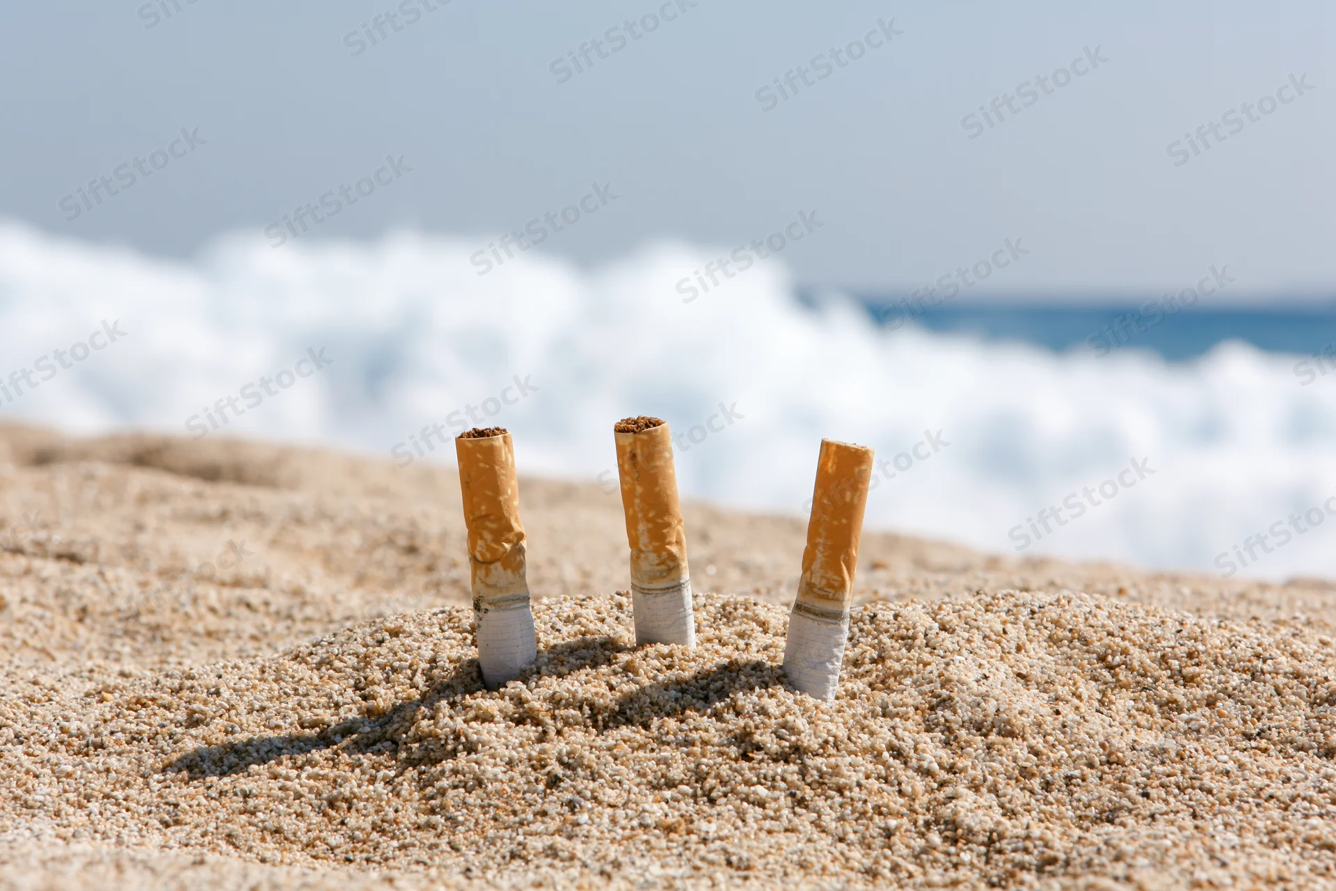 Three cigarette butts stuck upright in dry beach sand with ocean waves in background