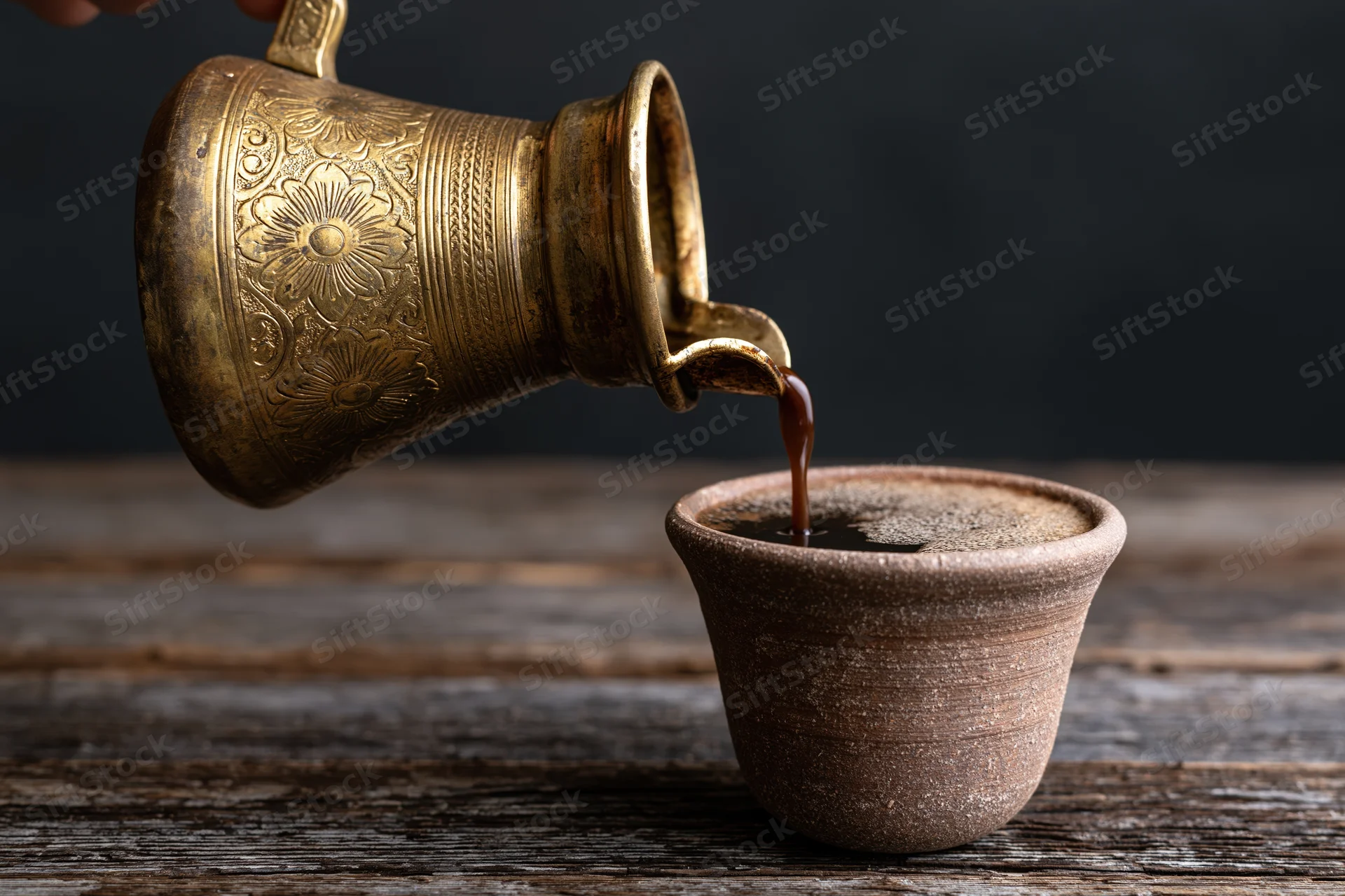 Turkish coffee being poured from an engraved brass cezve into a clay cup on a wooden table