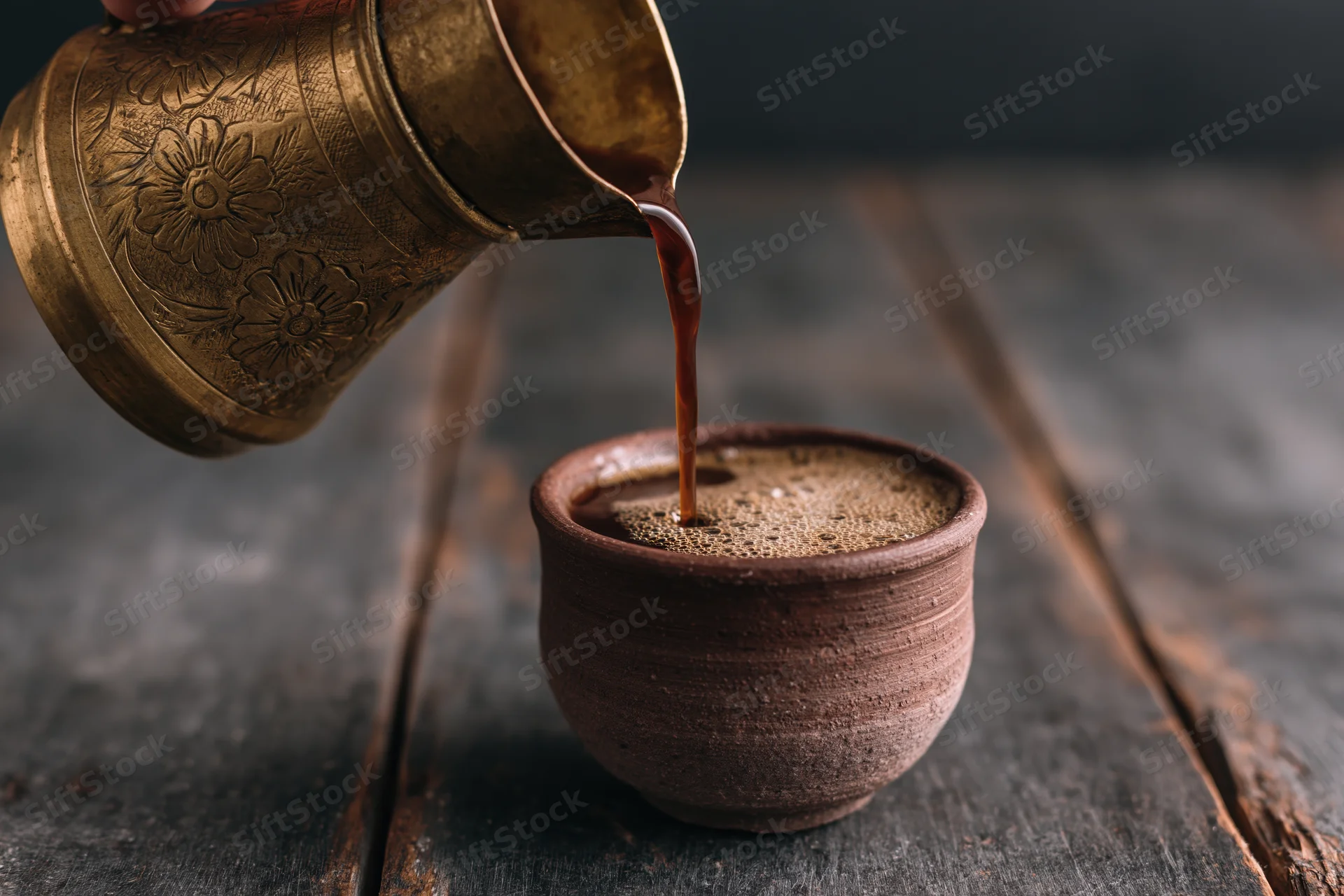 Turkish coffee being poured from an engraved brass cezve into a ceramic cup on a dark wooden table