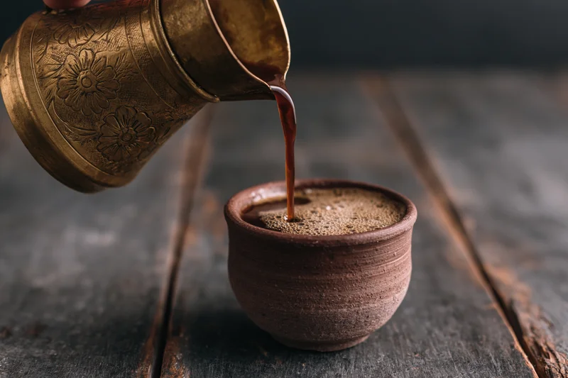 Turkish coffee being poured from an engraved brass cezve into a ceramic cup on a dark wooden table