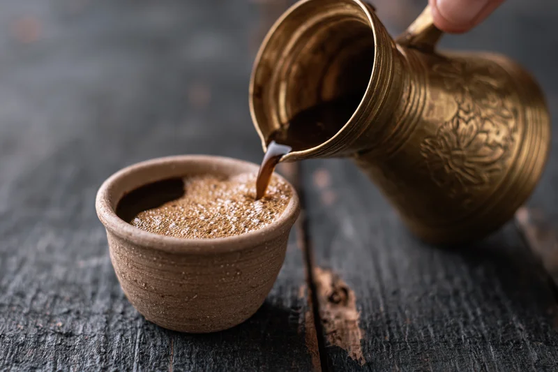 Turkish coffee being poured from a brass cezve into a ceramic cup on a dark wooden table